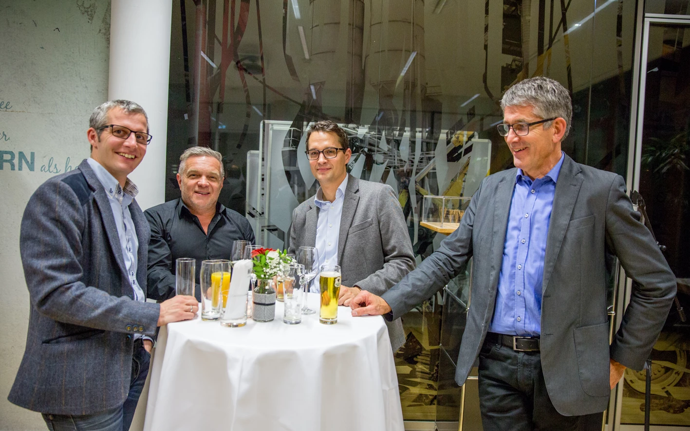 Vier Männer in Anzügen stehen lachend um einen Stehtisch mit Getränken bei einer Veranstaltung (Hubert, Geburtstag).