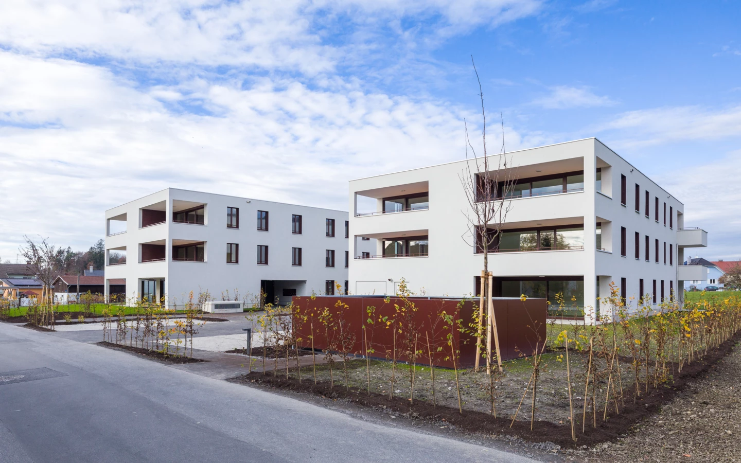 Zwei moderne, weiße Mehrfamilienhäuser mit Balkonen und Fenstern, umgeben von jung gepflanzten Bäumen und Sträuchern an der Unterhochstegstraße.