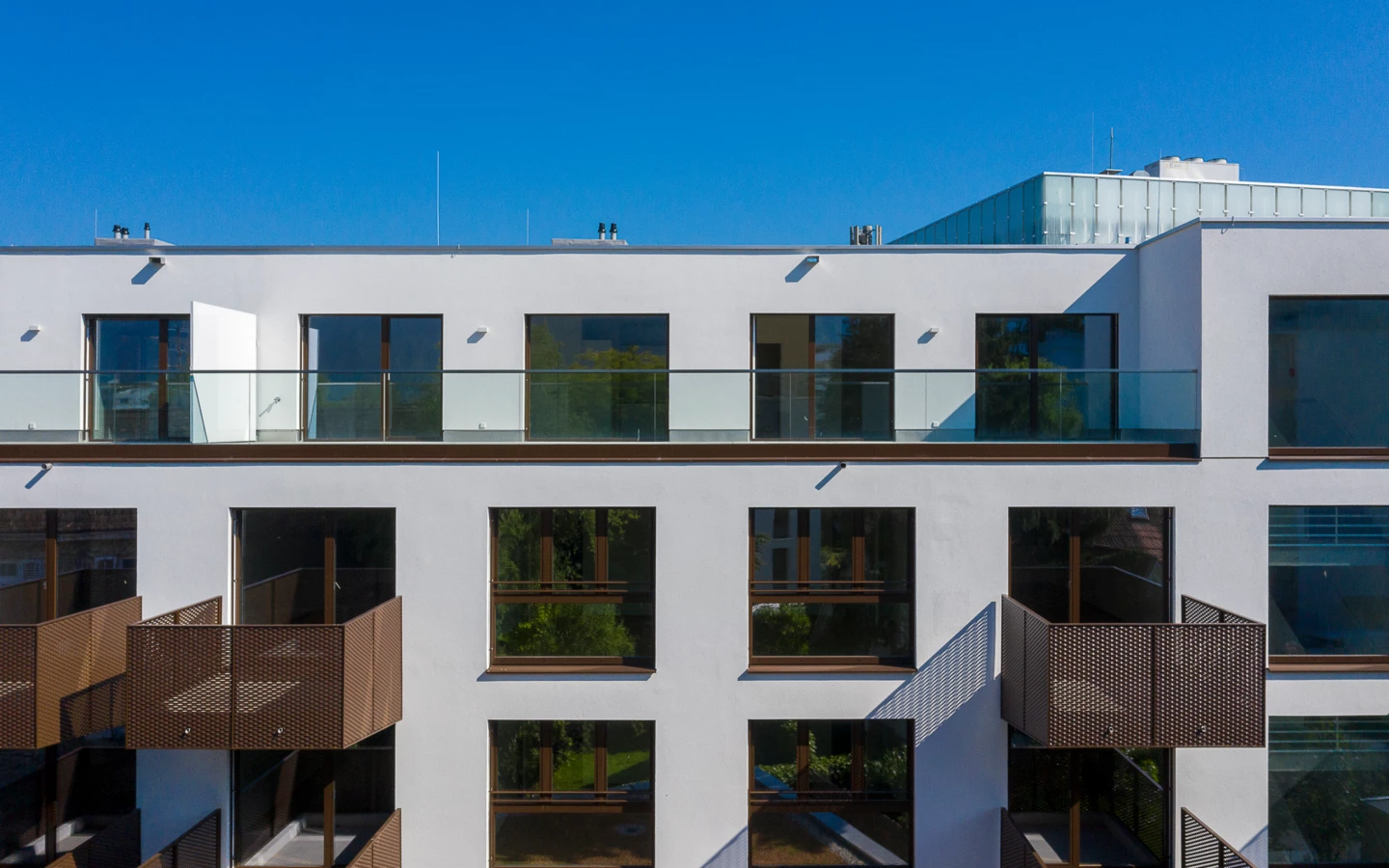 Fassade eines weißen Gebäudes mit braunen perforierten Metallbalkonen und großen Fenstern unter blauem Himmel (Dornbirn).