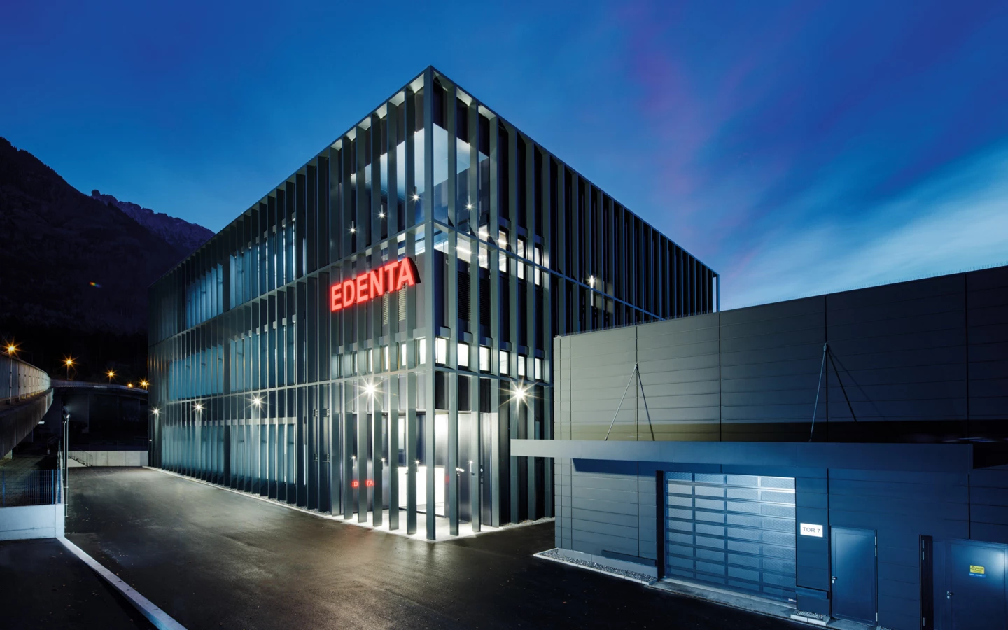 Modernes Geschäftsgebäude von Edenta bei Dämmerung mit beleuchtetem Logo und klarer Architektur unter blauem Himmel.