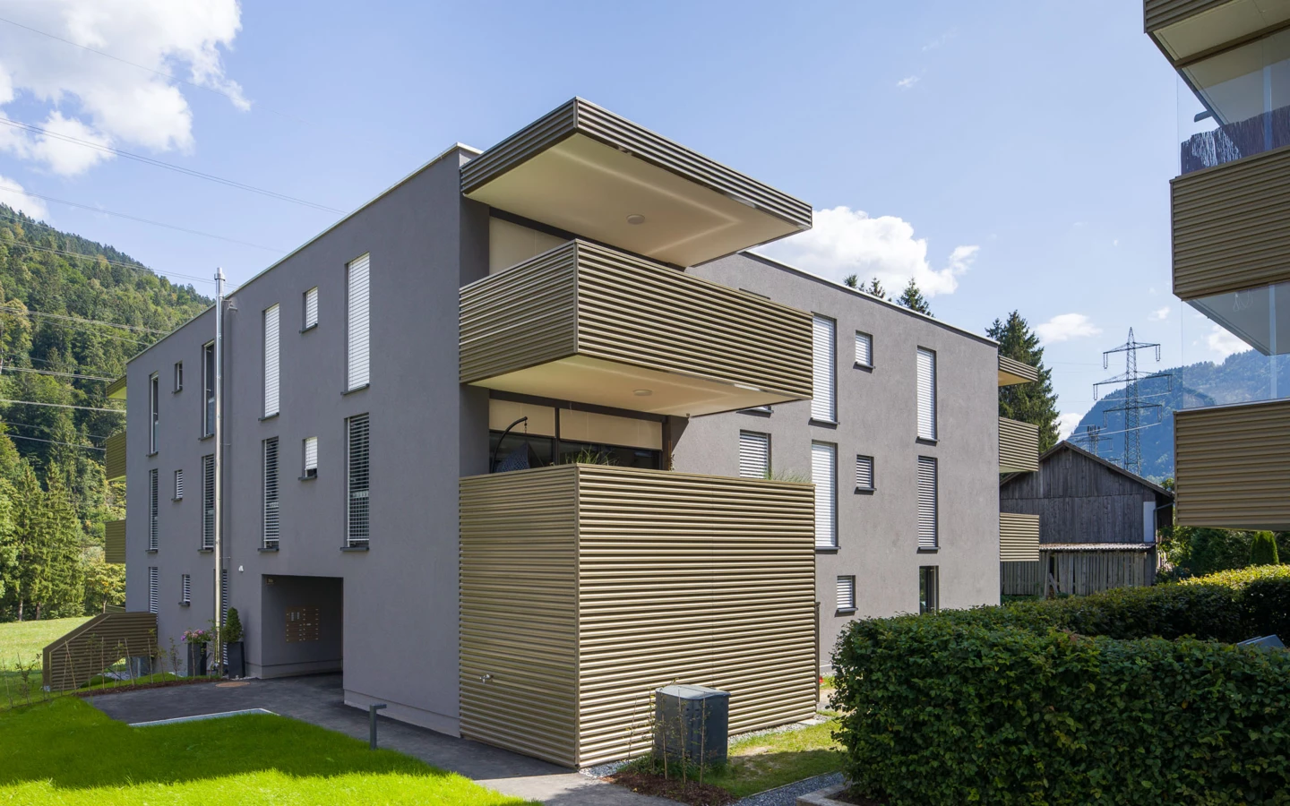 Modernes Wohngebäude in der Bahnhofstraße Vandans mit grauer Fassade, Balkonen mit Holzlamellen und Grünflächen bei Tageslicht (Wohnanlage, Bahnhofsstrasse, Übersicht).