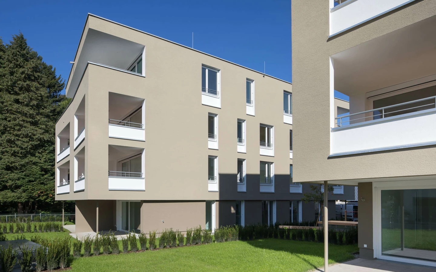 Zwei moderne helle Wohnanlagen Widum Lustenau auf einer grünen Wiese mit Bäumen unter blauem Himmel.