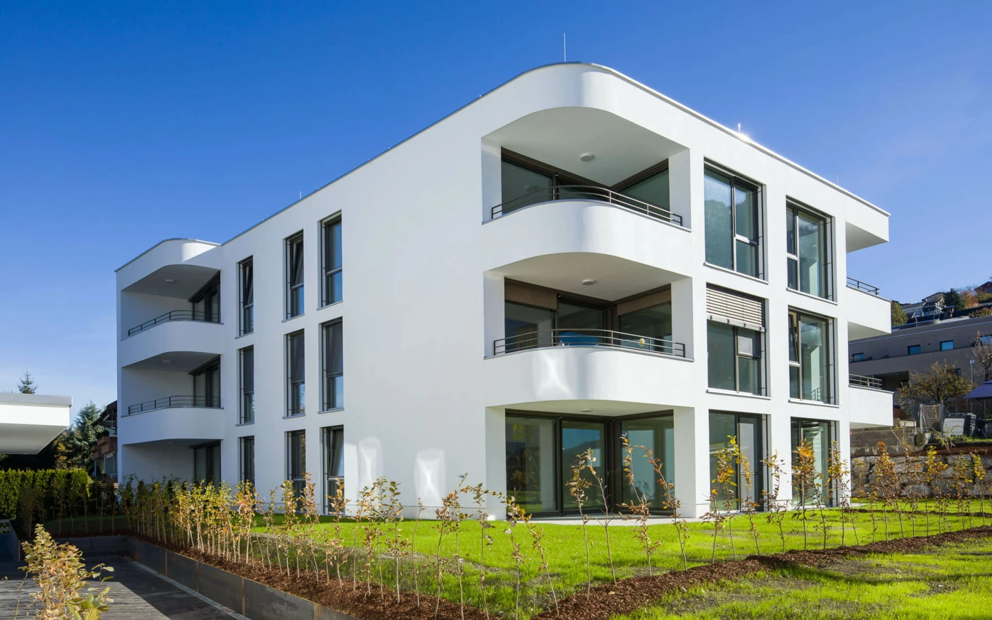 Moderne weiße Wohnanlage mit abgerundeten Balkonen, großen Fenstern, grünen Rasenflächen und jungen Bäumen unter blauem Himmel in Feldkirch (Wolf-Huber-Straße).