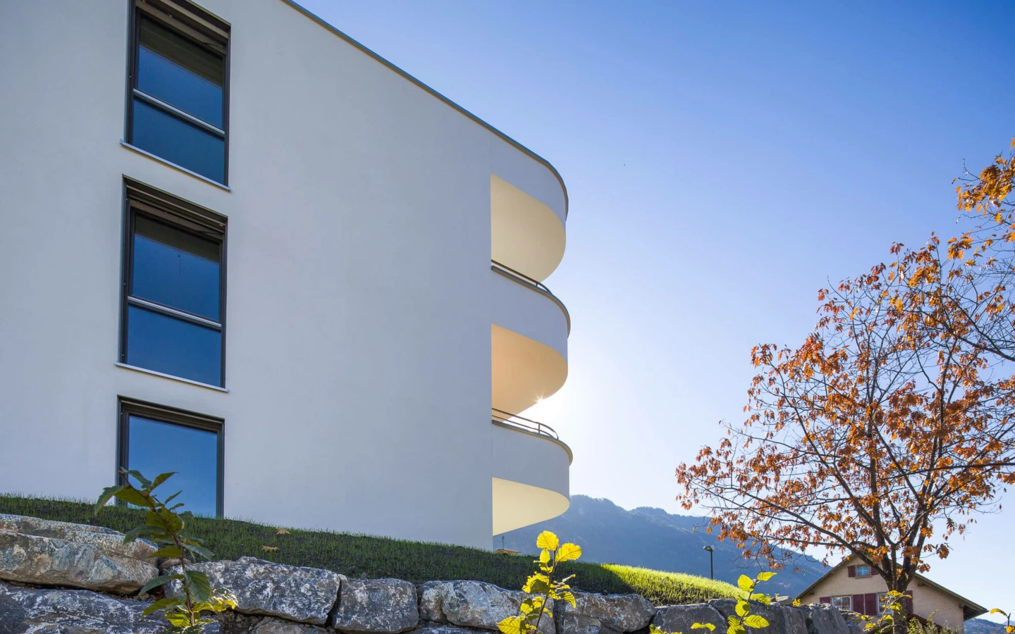 Modernes weißes Gebäude mit runden Balkonen und Fenstern über einer Steinmauer, Herbstbaum und Berge im Hintergrund unter blauem Himmel in Feldkirch (Wolf-Huber-Straße, Wohnanlage).