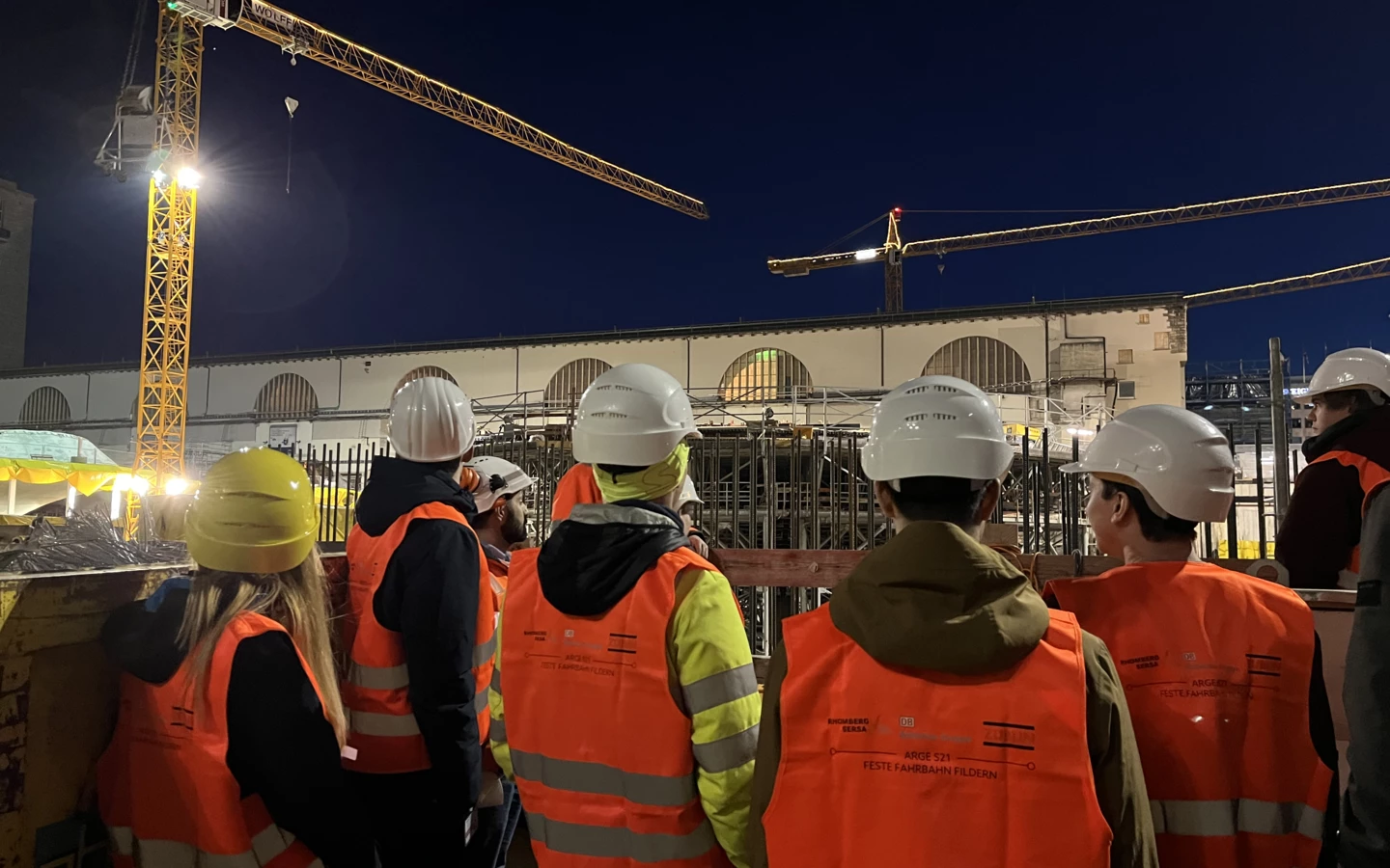 Arbeiter in Warnwesten und Helmen betrachten bei Nacht eine Baustelle mit Kranen und Gebäuden in Fildern (Rankweil, Deutschland, Kran).