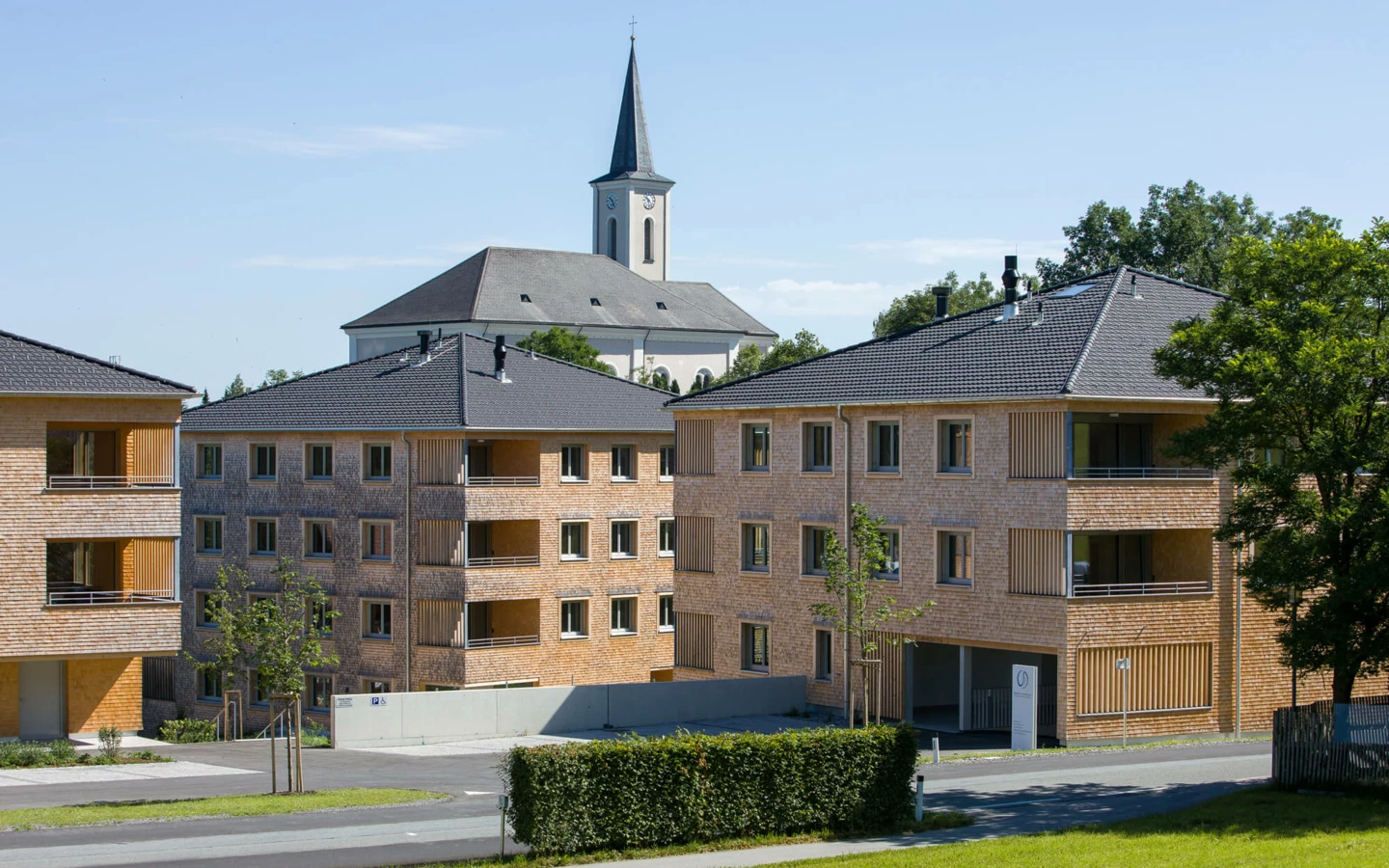 Zwei moderne Mehrfamilienhäuser mit Holzfassade und grauen Dächern in Alberschwende, mit Kirche im Hintergrund (Wohnanlage).