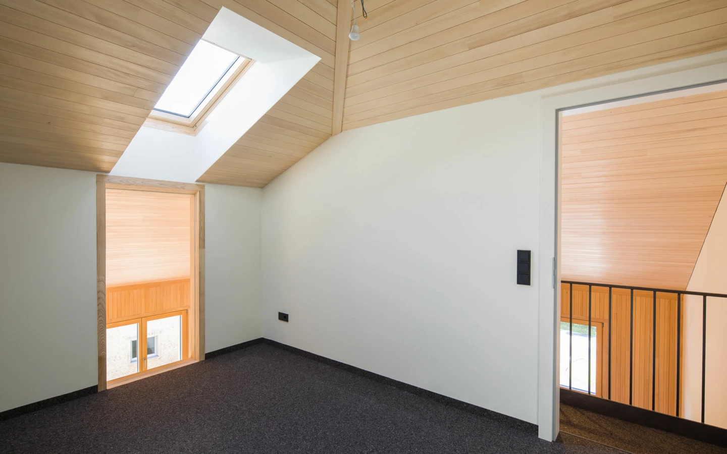 Dachgeschosszimmer mit Holzbalkendecke, schrägem Dachfenster, modernem Teppichboden und Fenster zum Treppenhaus. Eine Glühbirne hängt an der Decke (Wohnanlage, Alberschwende).