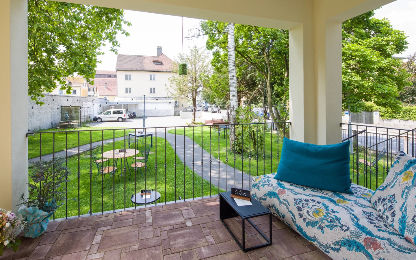 Blick von einer Terrasse mit gemusterter Liege und türkisfarbenem Kissen auf einen begrünten Garten mit Café-Tischen und einem Weg (Wunderkind, Backstube).