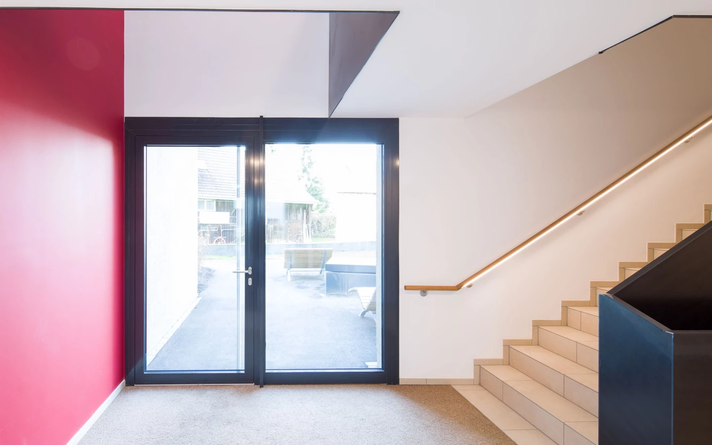Eingangsbereich mit Glastür zu einem Außenbereich, einer roten Wand, Treppen und einem beleuchteten Holzgeländer (Bäumleareal).