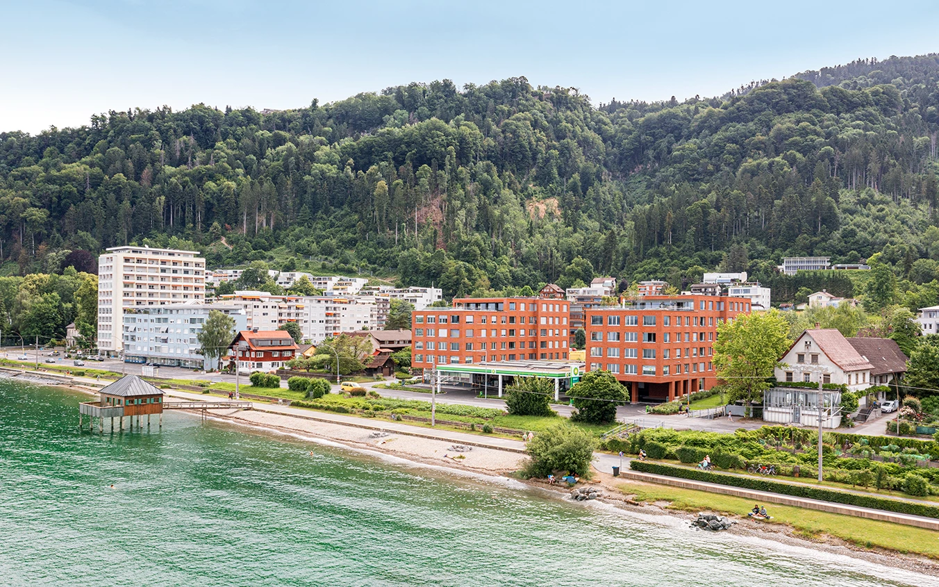 Mehrere Wohngebäude am Ufer des Bodensees in Lochau, darunter neue rote Gebäude und ältere weiße Türme, mit bewaldeten Hügeln im Hintergrund (Seeblick).