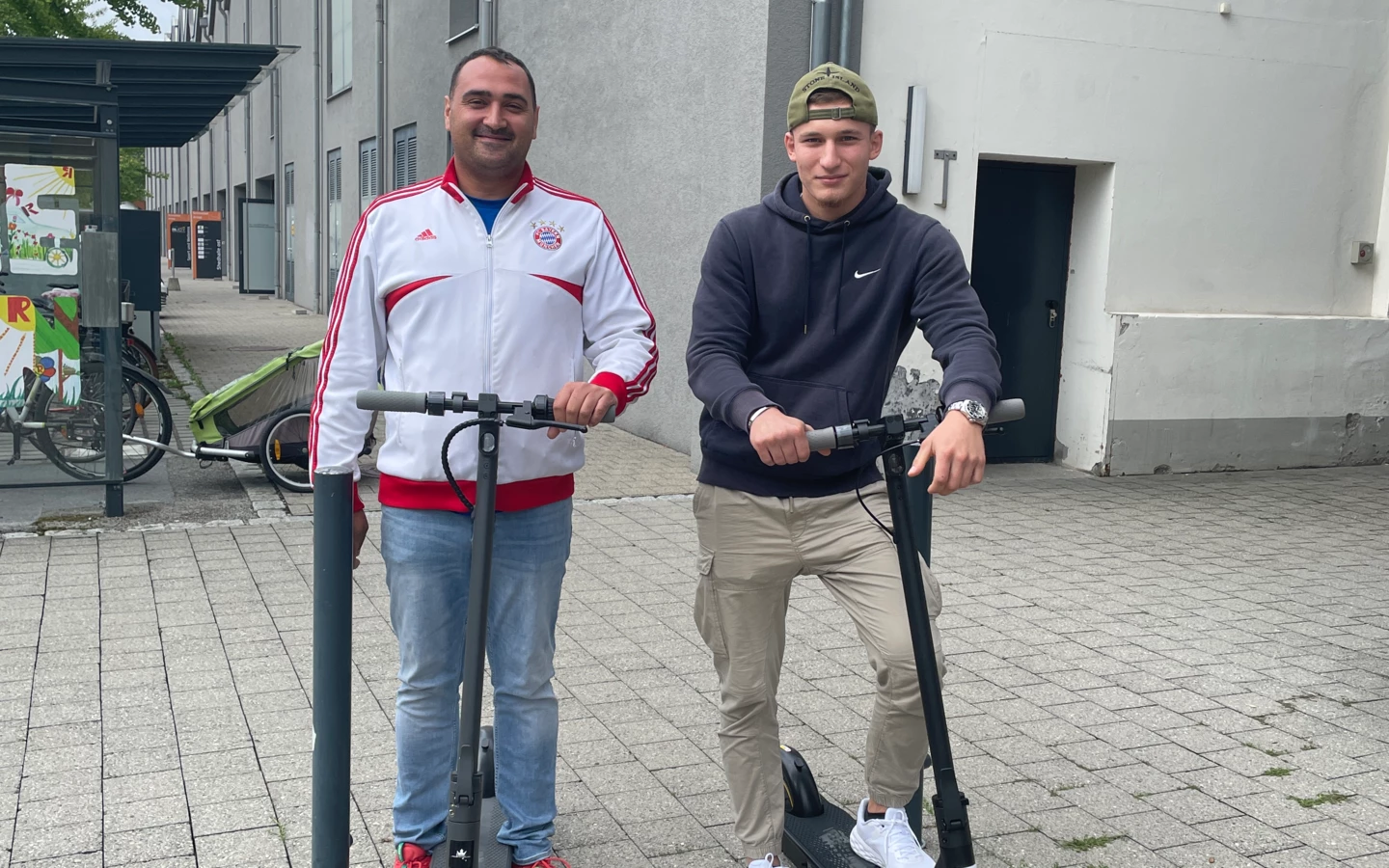 Zwei junge Männer stehen lächelnd mit schwarzen E-Scootern auf einem gepflasterten Gehweg vor modernen Gebäuden, links ein Fahrradständer (Lehre).