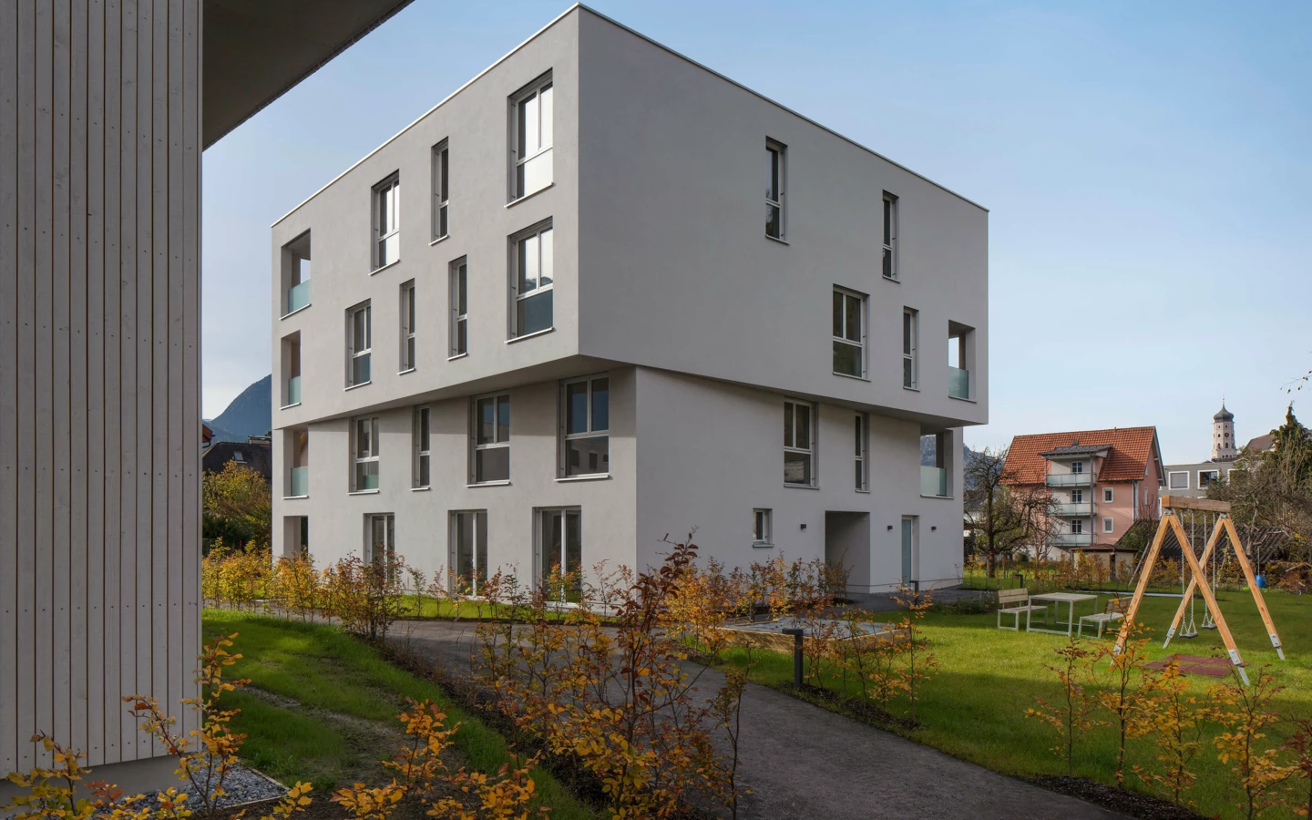 Moderne Wohnanlage Zürcherstrasse Bludenz mit weiß verputzten Gebäuden und Holzverkleidung, umgeben von Grünflächen und einem Spielplatz an einem sonnigen Tag.