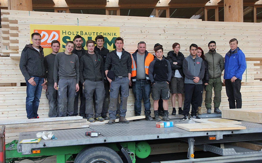 Eine Gruppe junger Männer und zwei ältere Männer posieren vor einem Stapel Holzbretter und einem Schild mit der Aufschrift "Holz Bautechnik Solm" (Lehrlingstag).