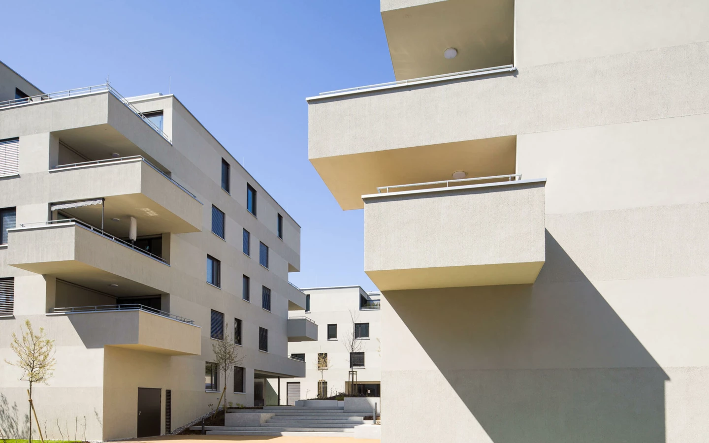 Moderne Wohnanlage mit beigen Fassaden unter blauem Himmel, mit Balkonen und dunklen Fenstern, dazu jung bepflanzte Bäume und Wege (Wuhrbaumweg).