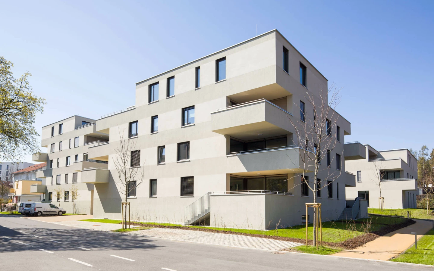 Moderne, hellgraue Wohngebäude mit Balkonen und großen Fenstern unter blauem Himmel, umgeben von jungem Grün (Wuhrbaumweg, Wohnanlage)