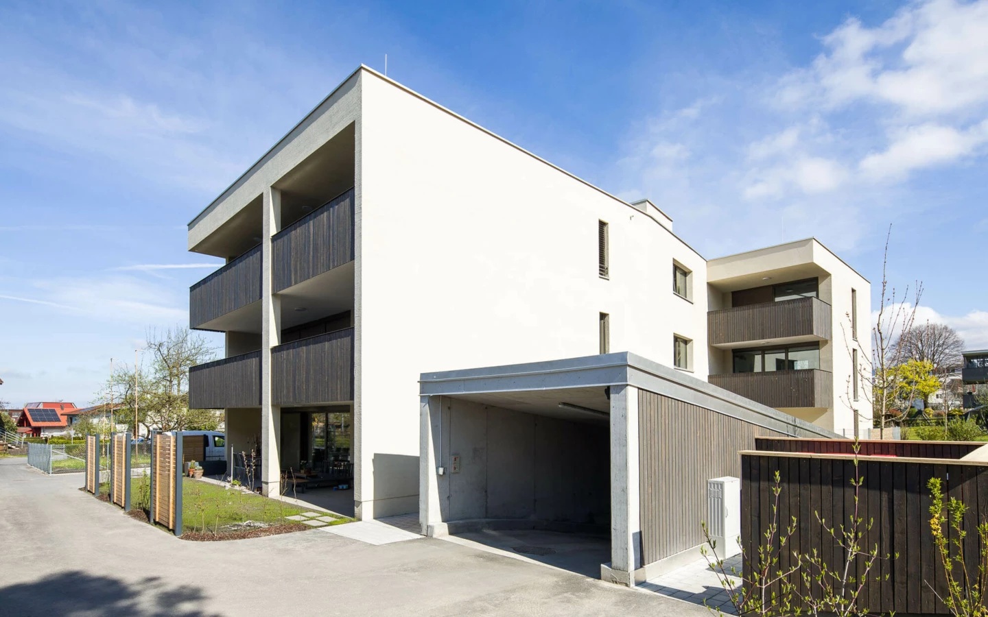Modernes Wohngebäude in der Feldgasse Dornbirn mit weißer Fassade, Holzbalustraden und einem offenen Carport unter blauem Himmel (Wohnanlage).