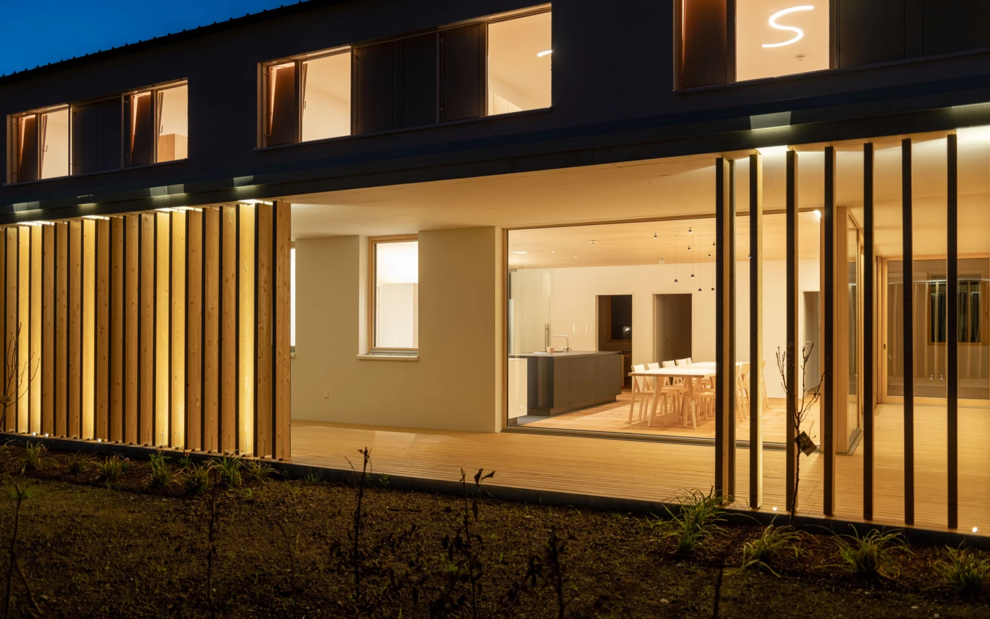 Modernes Haus bei Nacht mit beleuchteter Holzfassade, weit geöffneten Terrassen und warm beleuchteten Innenräumen, die ein Esszimmer und eine Küche zeigen (Wolfurt, Dieter).