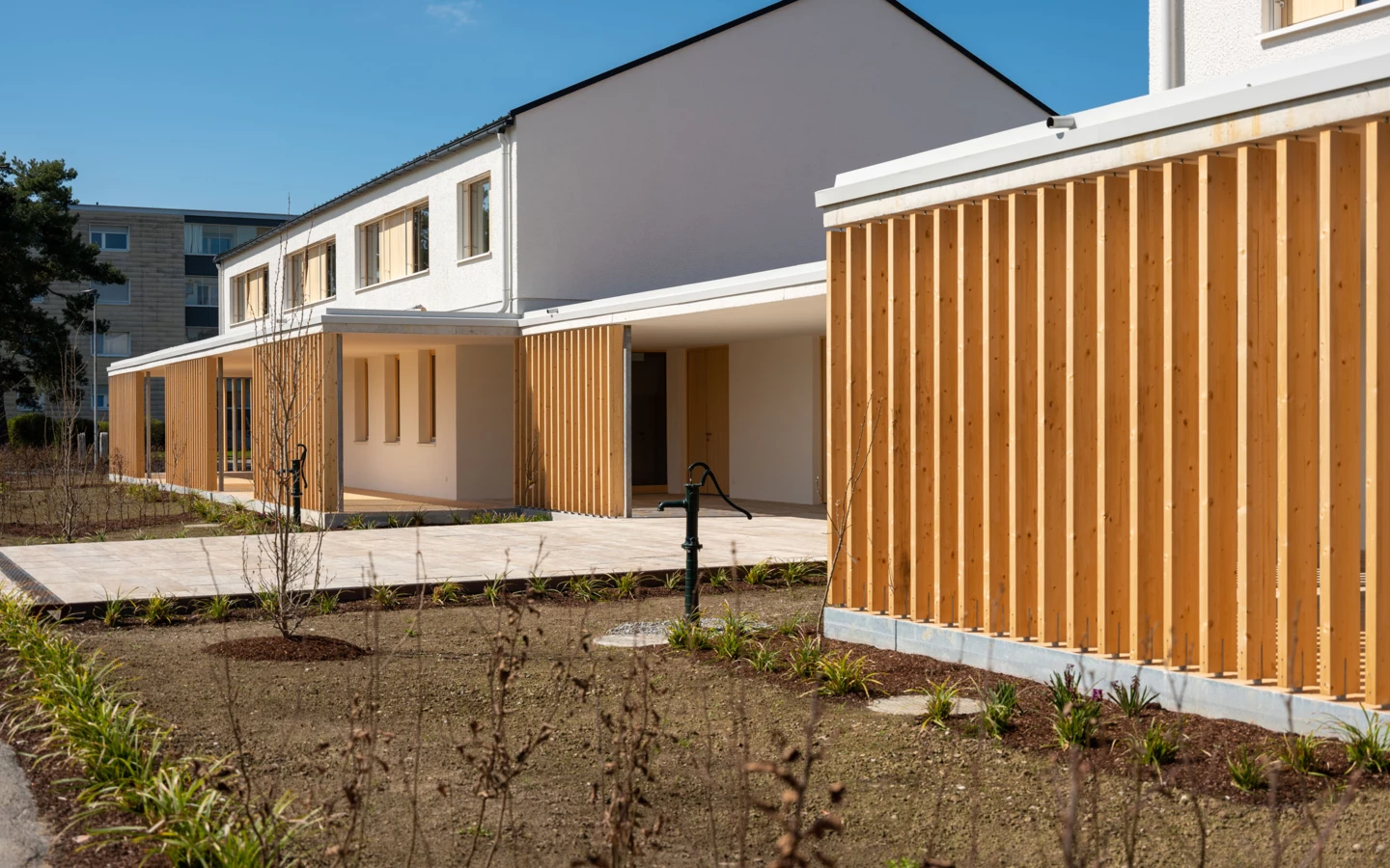 Außenansicht eines modernen weißen Gebäudes mit Holzlamellenfassade und angelegten Grünflächen unter blauem Himmel in Wolfurt (Dieter).