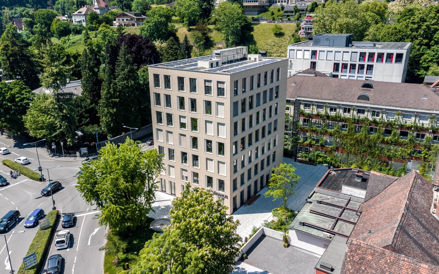 Aerial view of the modern Baerahus building with solar panels on the roof, surrounded by green trees, other buildings, and a multi-lane (Feldkirch)
