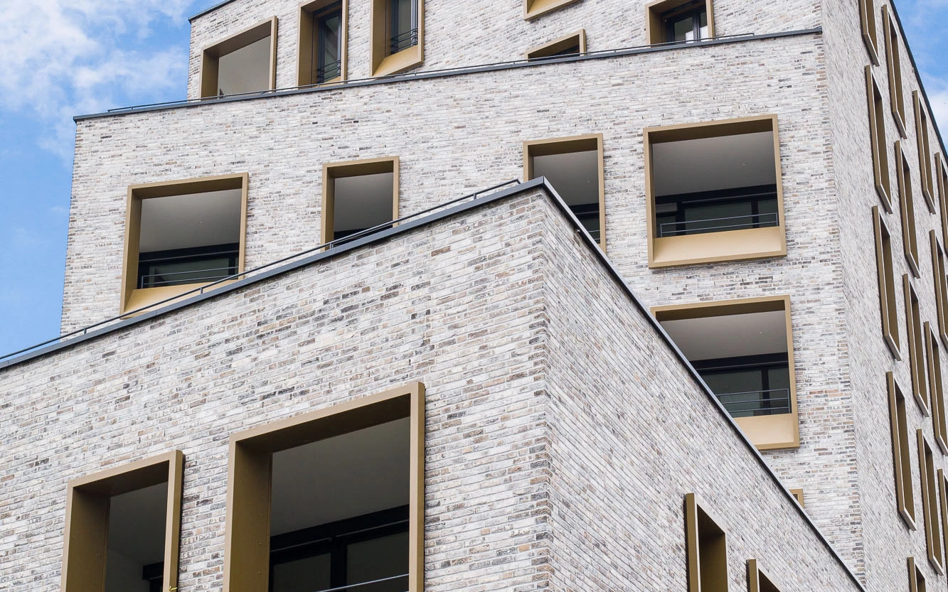 Modernes Gebäude in Dornbirn, Kreuzgasse 4, mit heller Ziegelfassade, eckigen, goldfarbenen Fensterrahmen und einem Baum im Vordergrund.