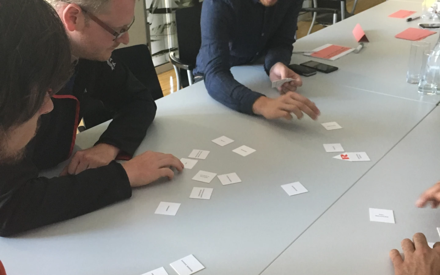 Drei Männer sitzen an einem Tisch und verschieben kleine Kärtchen, die mit Text bedruckt sind, in einem Workshop.