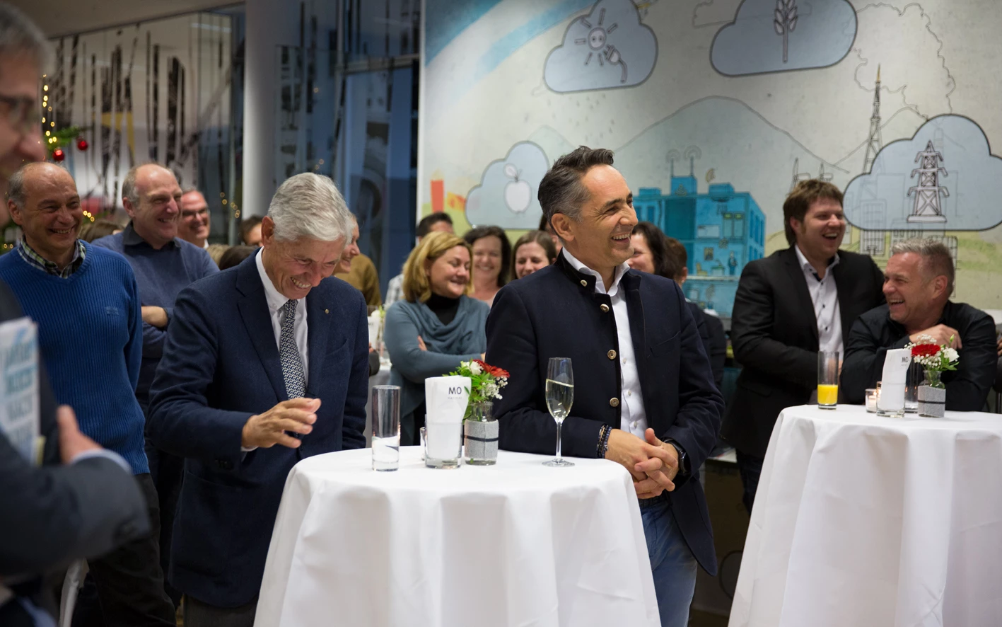 Eine Gruppe lachender, festlich gekleideter Männer und Frauen steht in einem Raum an Stehtischen mit Getränken (Hubert, Geburtstag).
