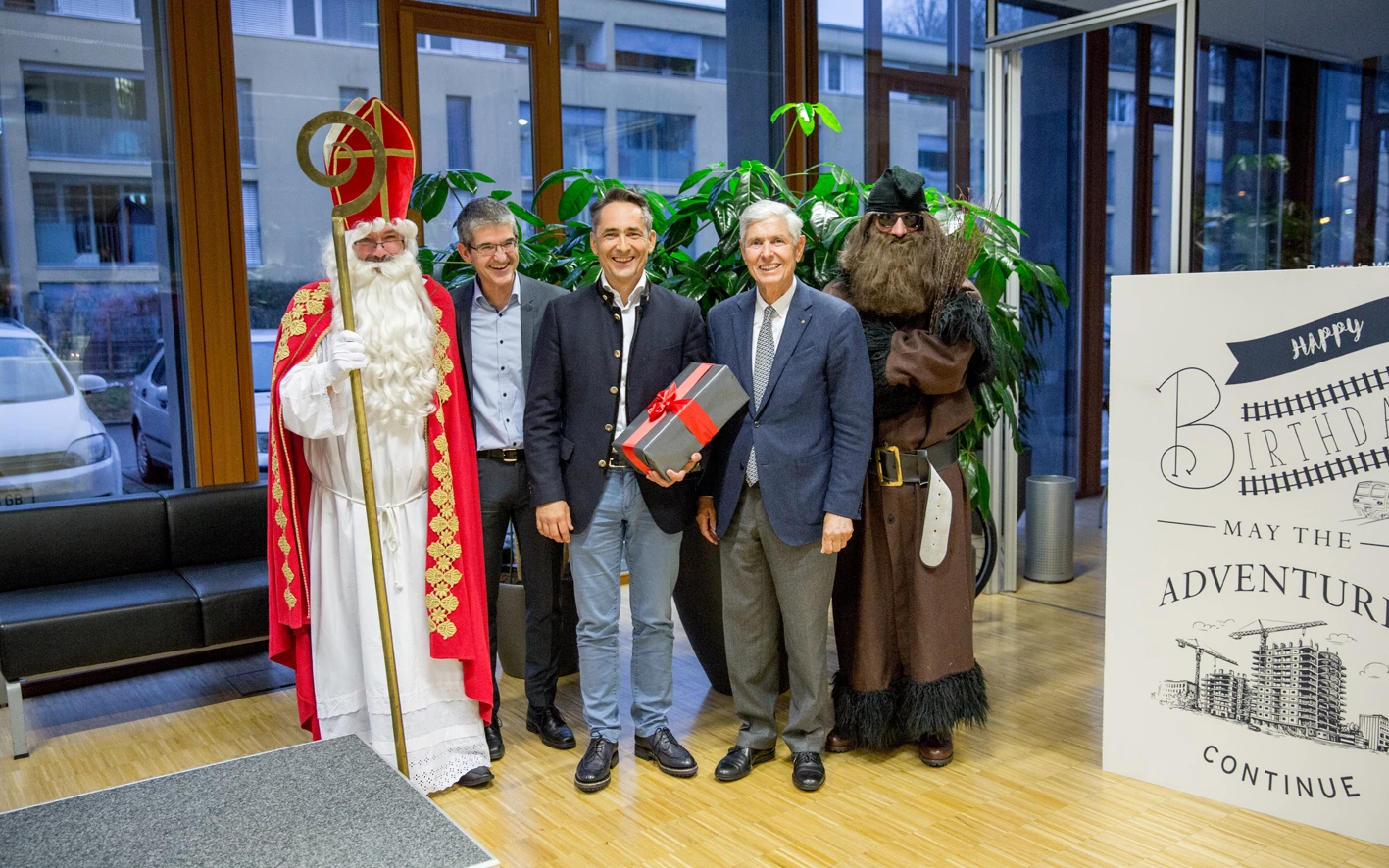 Vier Männer in Anzügen und Kostümen, darunter St. Nikolaus, stehen vor einer Zimmerpflanze und einem Geburtstagsplakat mit Bauvisualisierung (Hubert).