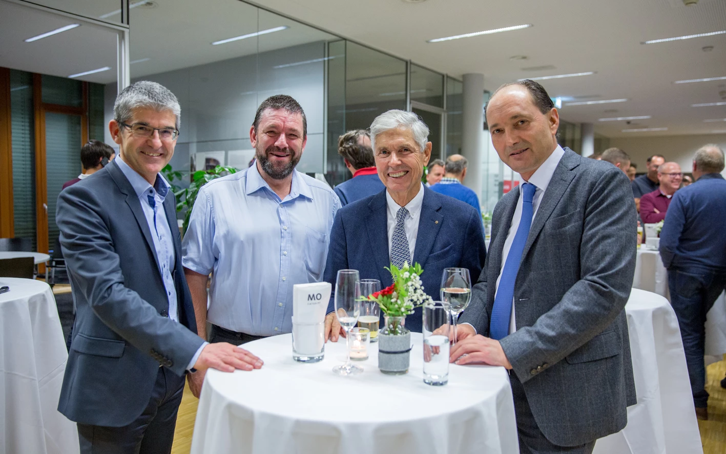 Vier lächelnde Männer in stilvoller Kleidung, die bei einer Veranstaltung an einem Stehtisch mit Getränken und Blumen stehen (Hubert, Geburtstag).