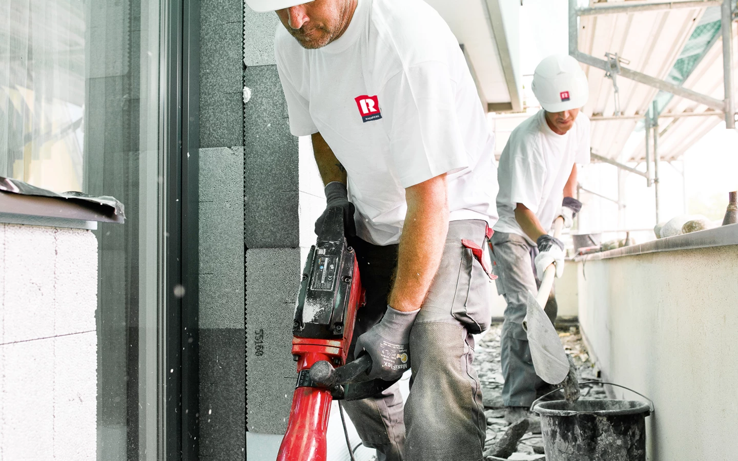 Zwei Bauarbeiter in Schutzkleidung auf einer Baustelle. Einer verwendet einen Bohrhammer, der andere schaufelt Schutt in einen Eimer (Hochbau).