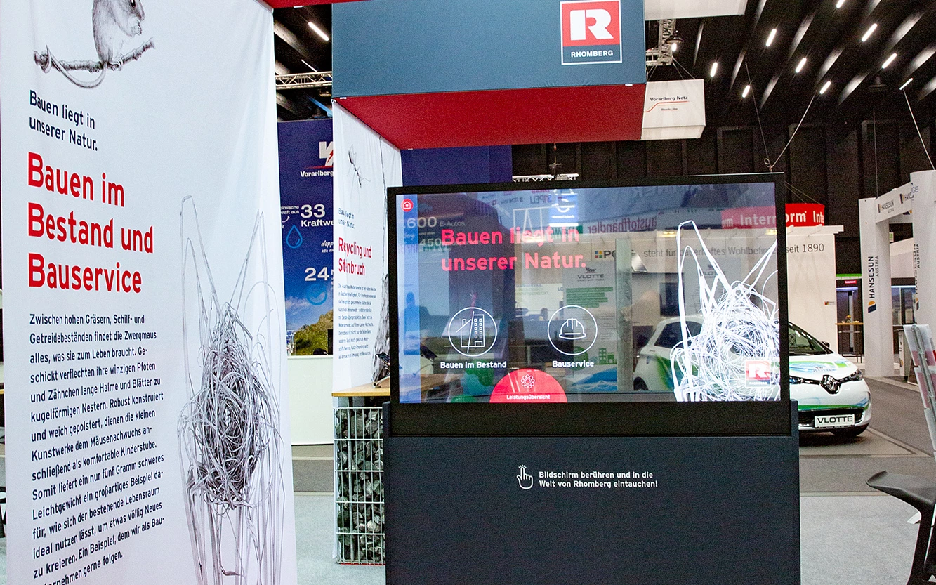 Rhomberg Bau Messestand mit interaktivem Bildschirm, rotem Teppich und Banner mit Mausmotiv und Text "Bauen liegt in unserer Natur"