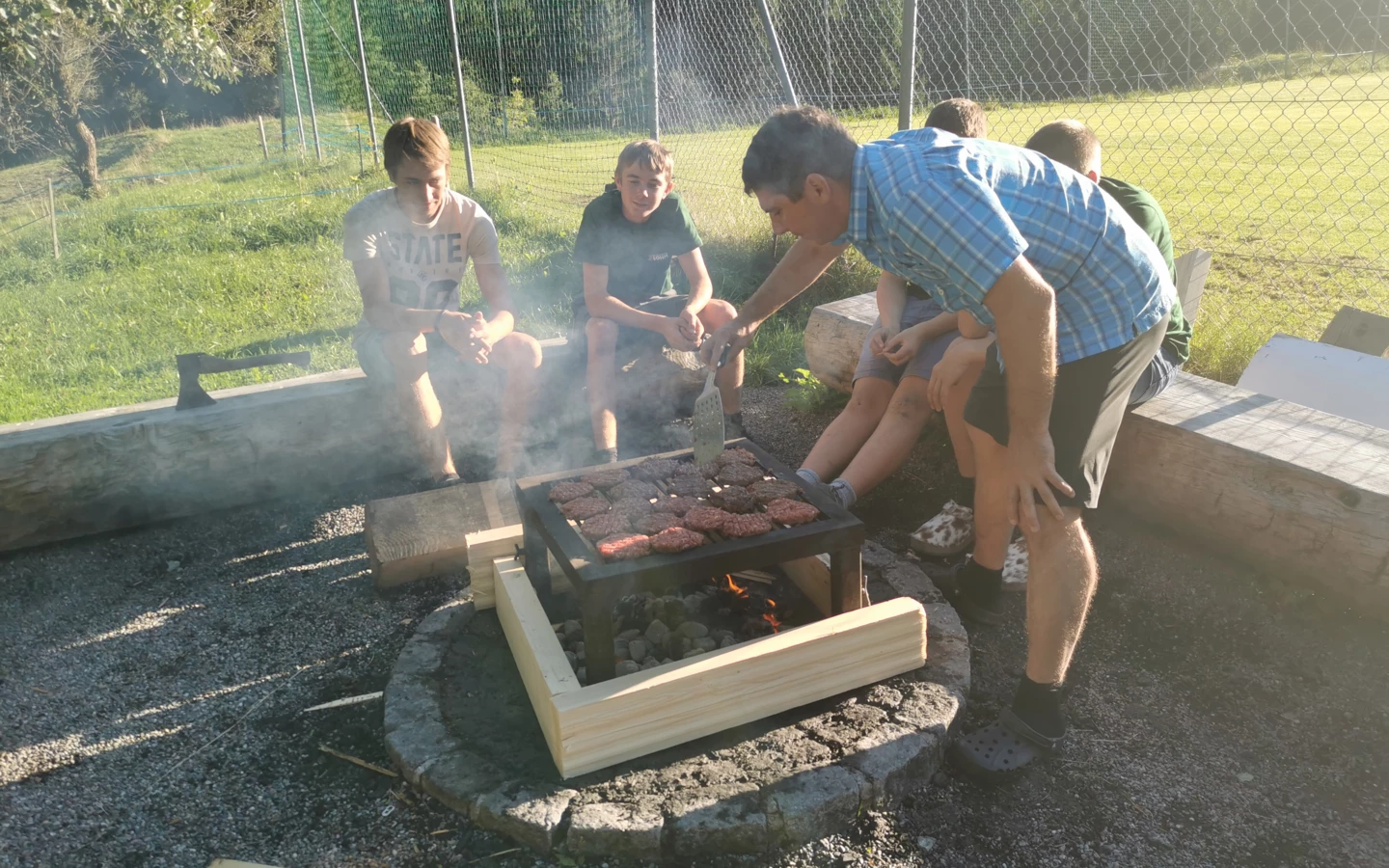 Ein Mann im blau karierten Hemd grillt Burger für eine Gruppe sitzender Jugendlicher beim Lehrlingscamp 2019.