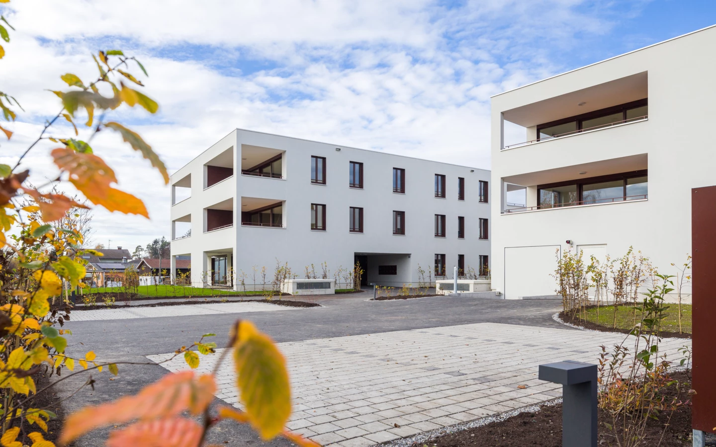 Zwei moderne weiße Mehrfamilienhäuser mit Balkonen und dunklen Fenstern, umgeben von gepflasterten Flächen und junger Bepflanzung unter blauem Himmel mit Wolken (Unterhochstegstrasse).