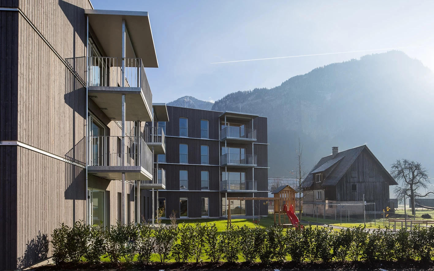 Moderne Wohngebäude mit Holzfassaden und Balkonen neben einem Spielplatz, einer Scheune und Bergen bei Sonnenlicht (Froschweg).