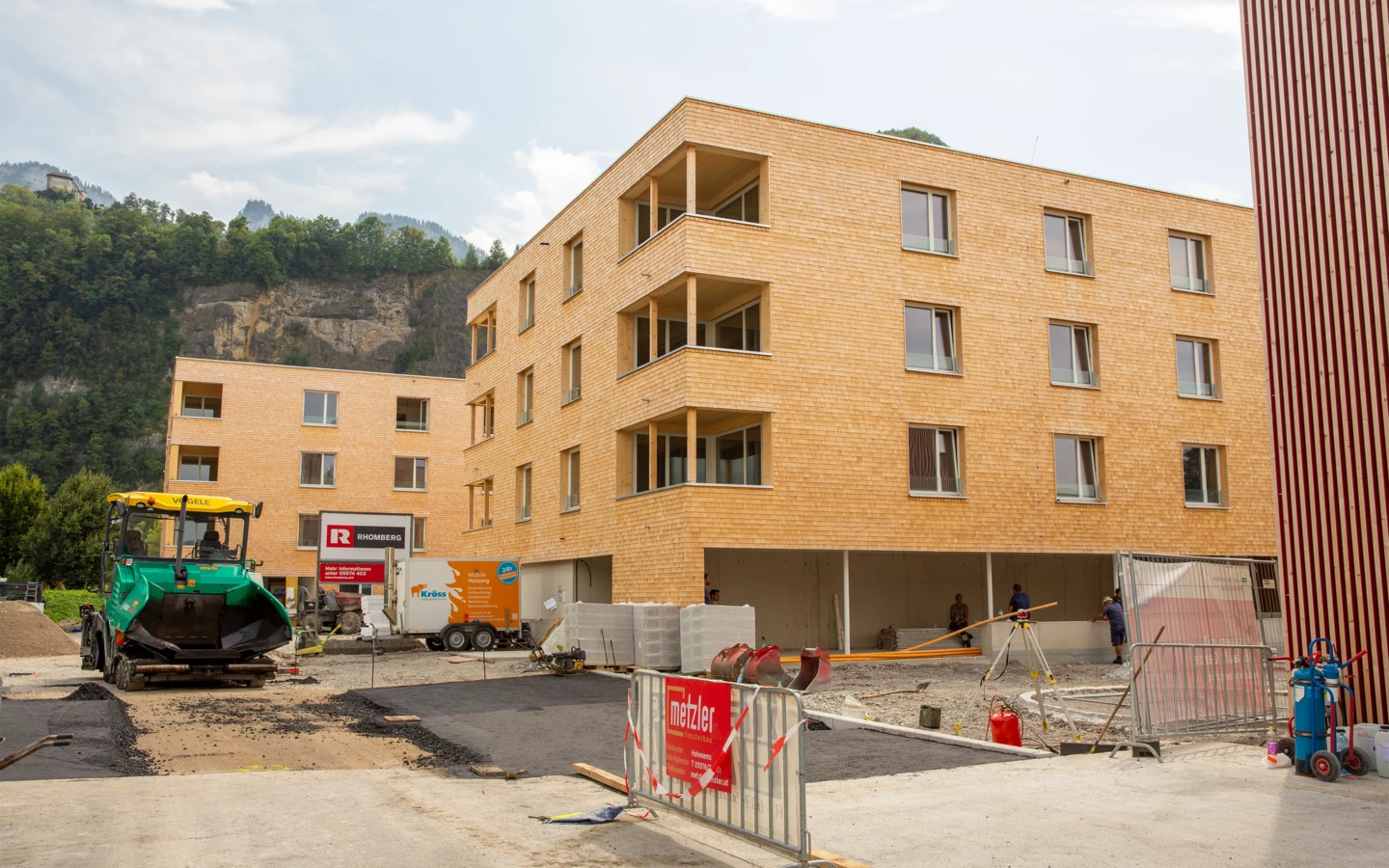 Zwei moderne Holzgebäude auf einer Baustelle mit einem grünen Asphaltfertiger und Bauarbeitern (Hellbrunnenstraße).