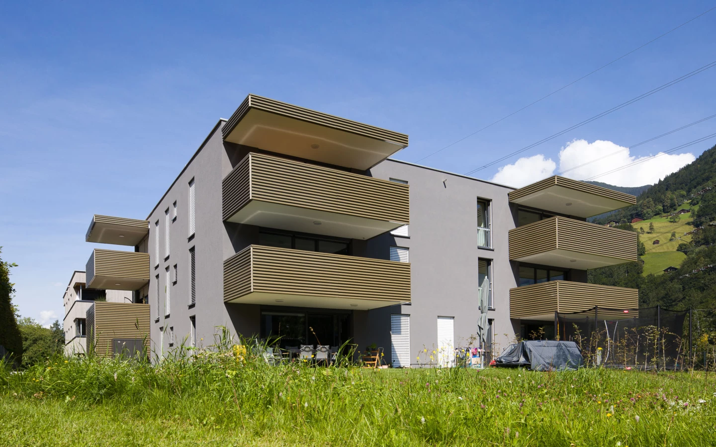 Moderne Wohnanlage in Vandans mit grauer Fassade, mehreren Balkonen mit Holzlamellen und einer grünen Wiese im Vordergrund unter blauem Himmel (Bahnhofstraße, Bahnhofsstrasse).