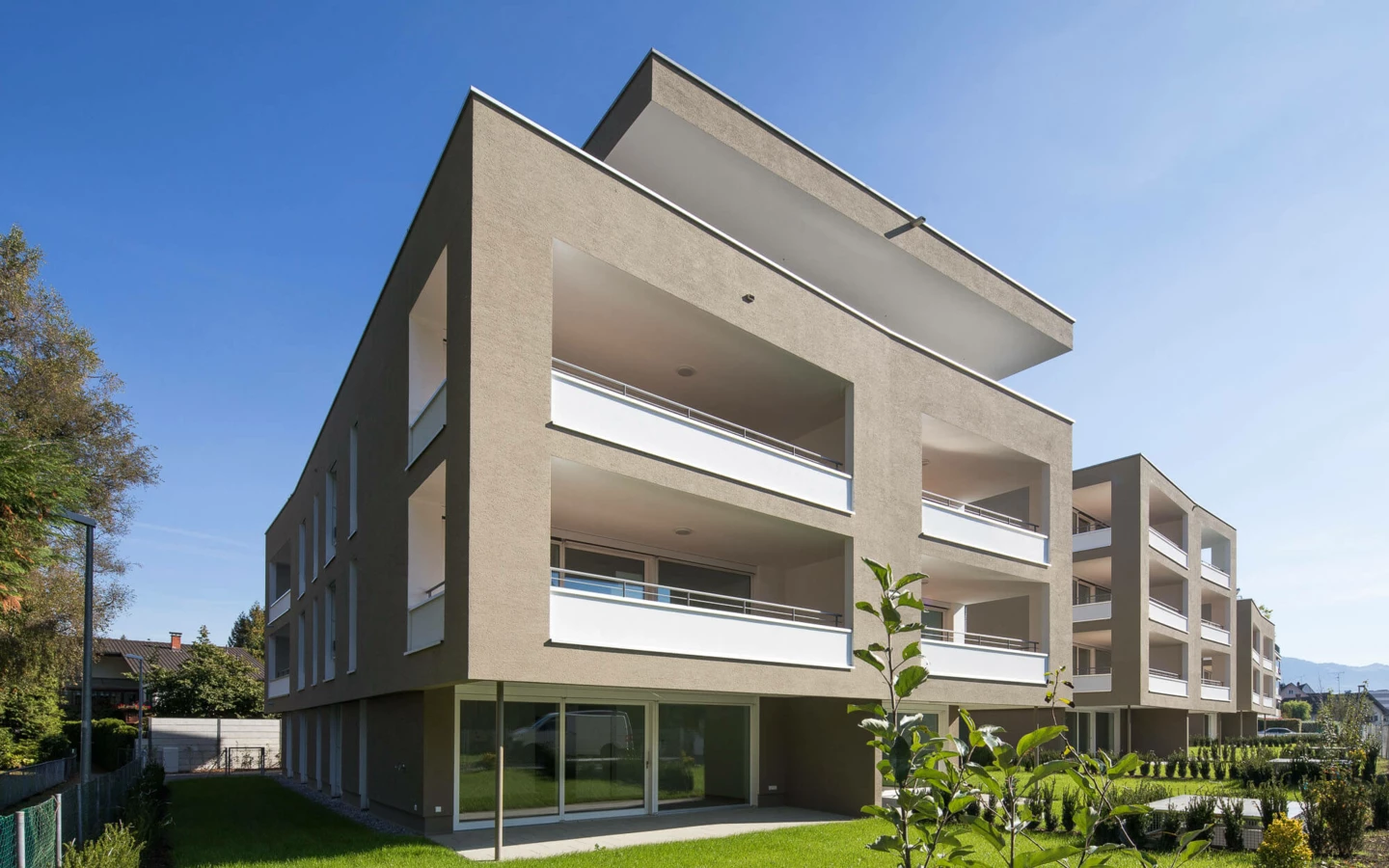 Moderne Wohngebäude der Wohnanlage Widum in Lustenau mit brauner Fassade, weißen Balkonen und gepflegtem Rasen unter blauem Himmel.