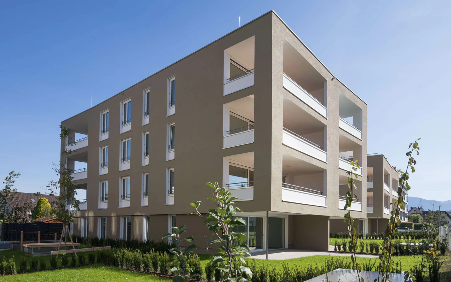 Moderne Wohnanlage Widum in Lustenau mit beigefarbenen Fassaden, weißen Balkonen und Grünflächen unter blauem Himmel.