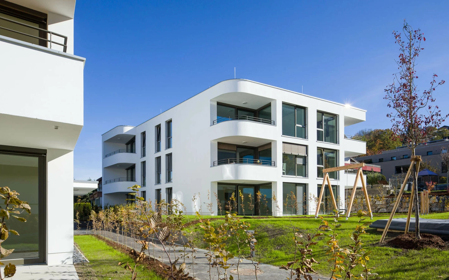 Moderne weiße Wohnanlage in Feldkirch mit gerundeten Balkonen, großen Fenstern, gepflegtem Rasen und einem Holzschaukelgerüst unter blauem Himmel (Wolf-Huber-Straße).