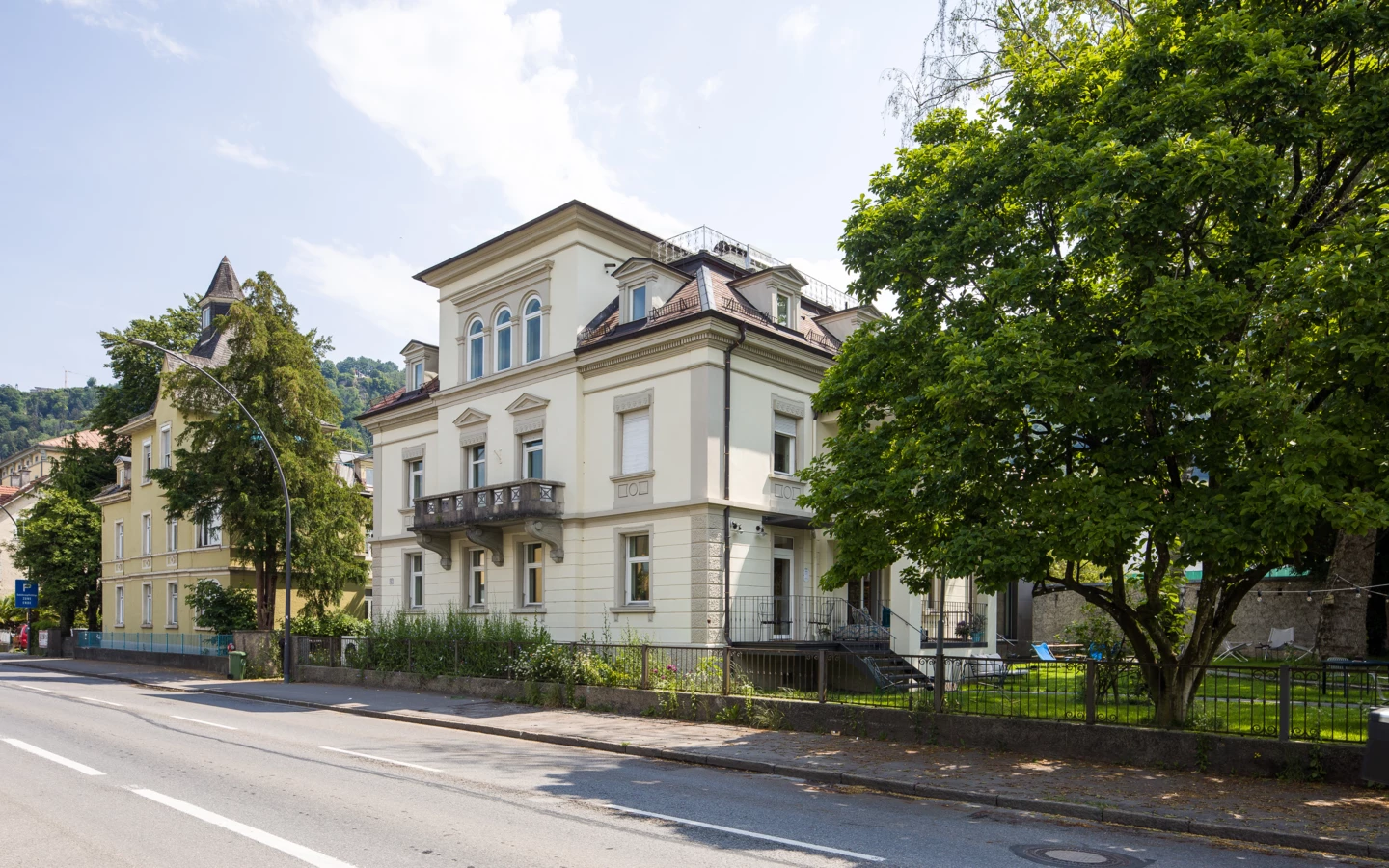 Eine Straße mit einem großen, cremefarbenen Gebäude mit Balkonen und einem hohen Baum daneben (Wunderkind, Backstube).