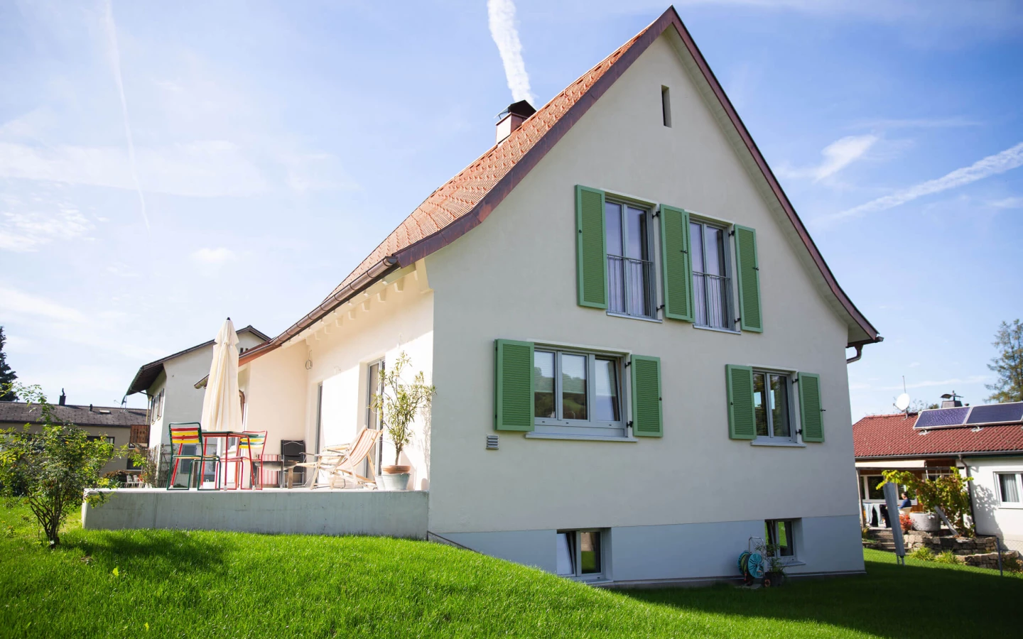 Einfamilienhaus in Dornbirn mit hellbeiger Fassade, terracottafarbenem Dach, grünen Fensterläden und einer Terrasse mit Gartenmöbeln (Generalsanierung).