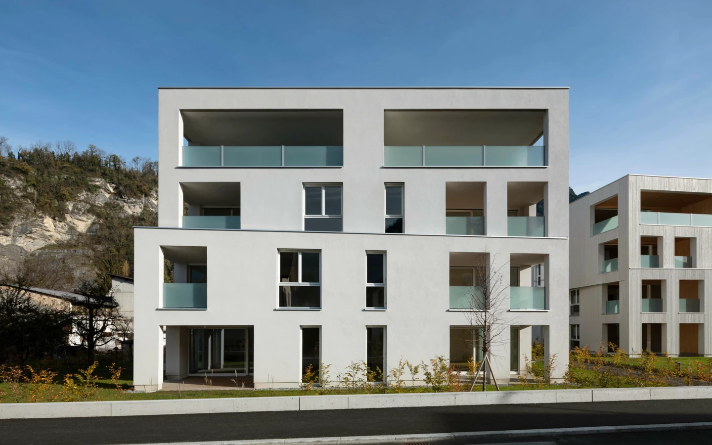Moderne Wohnanlage Zürcherstrasse Bludenz mit weißer Fassade, Glasbalkonen und Holzakzenten, vor bewaldetem Hang (Frontal).