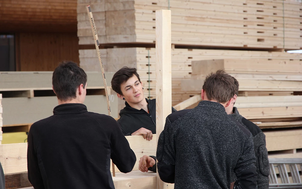 Drei junge Männer arbeiten mit Holzplatten und -balken in einer Halle, umgeben von gestapeltem Holzmaterial. Einer der Männer hält ein Maßband in der Hand (Lehrlingstag).