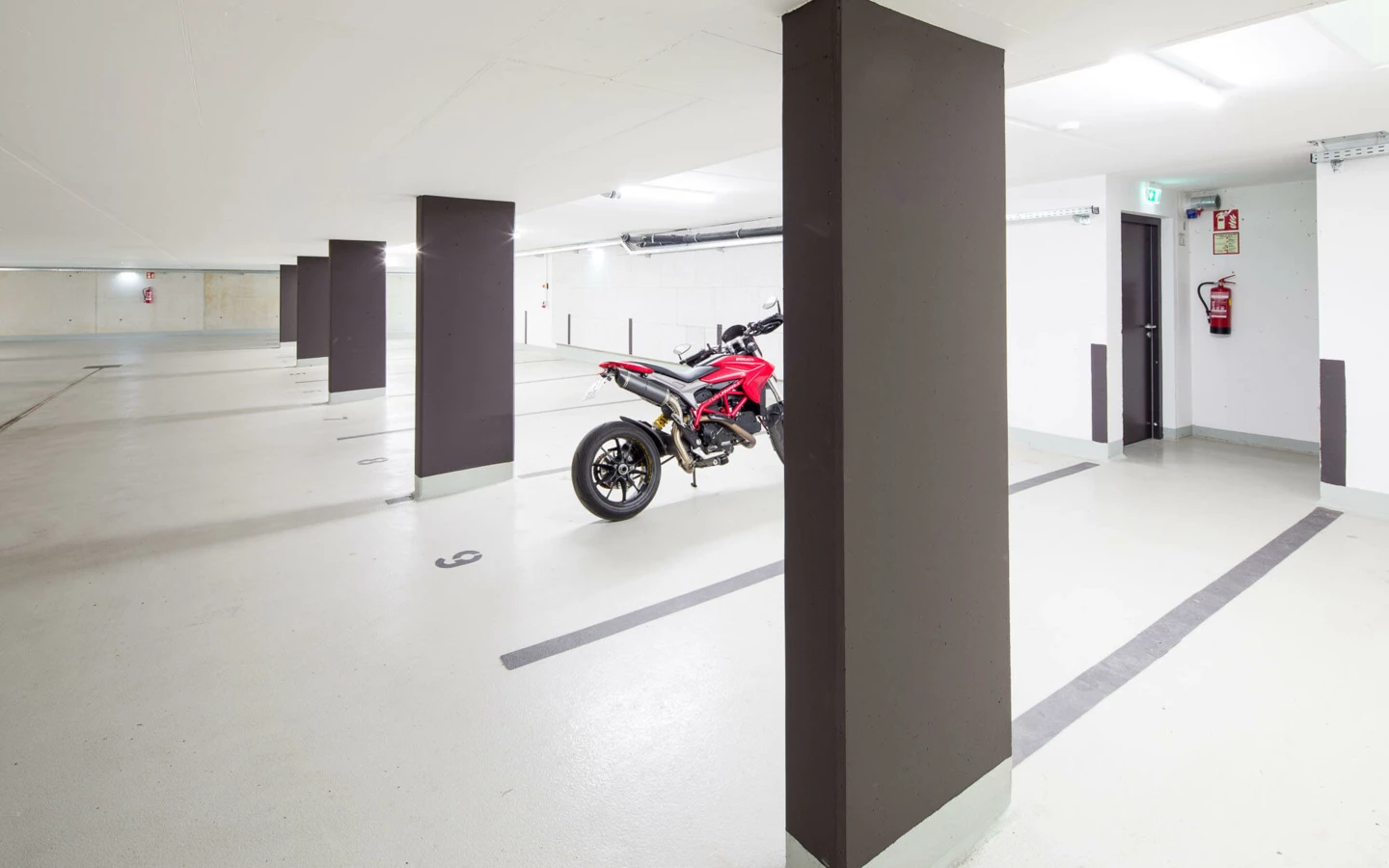 Eine leere, helle Tiefgarage mit Betonpfeilern und einem roten geparkten Motorrad (Wuhrbaumweg, Wohnanlage).