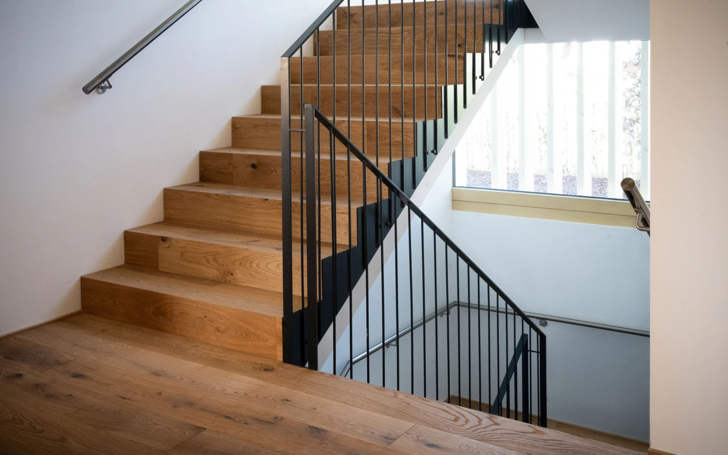 Eine moderne Holztreppe mit schwarzen Metallgeländern in einem Gebäude mit einem großen Fenster seitlich (Wolfurt, Dieter)