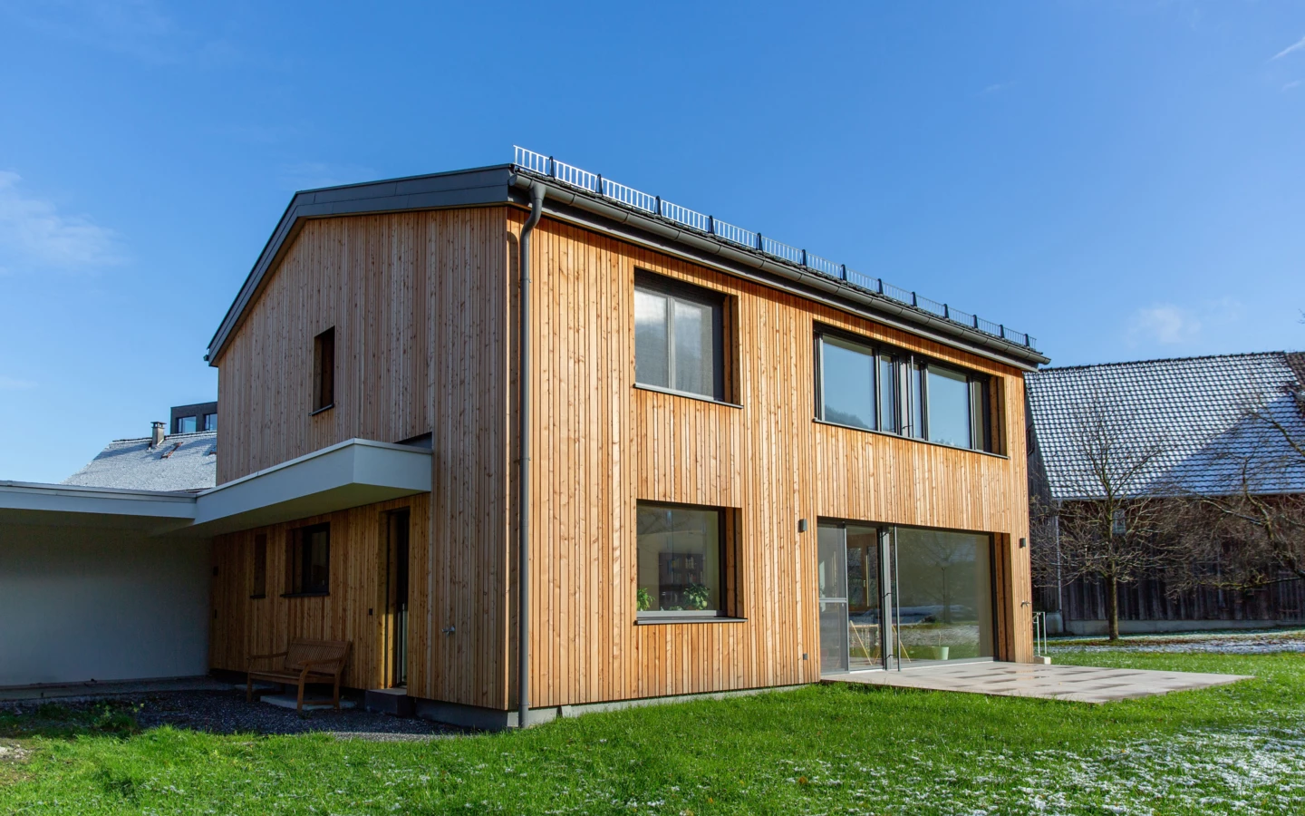 Modernes Einfamilienhaus in Dornbirn mit Holzfassade und großer Fensterfront auf einem leicht schneebedeckten Rasen unter blauem Himmel (Generalsanierung, Holzoptik).
