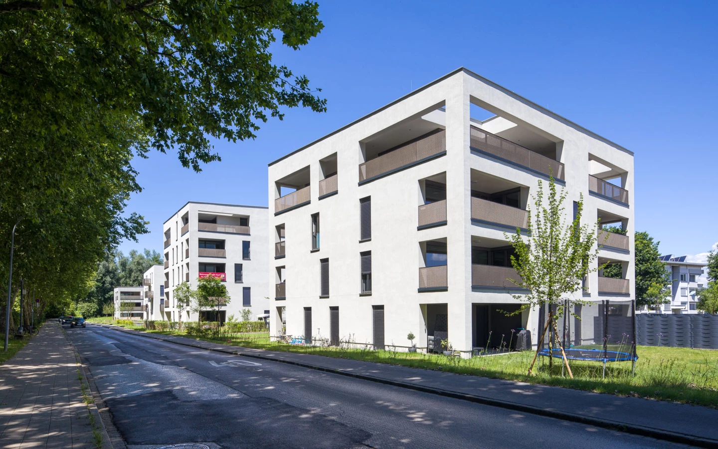Moderne weiße Wohngebäude mit Balkonen, umgeben von grünen Bäumen und Rasenflächen an einer Straße unter blauem Himmel. Ein Trampolin steht im Garten (Wohnanlage, Eigentumswohnung, Furt).