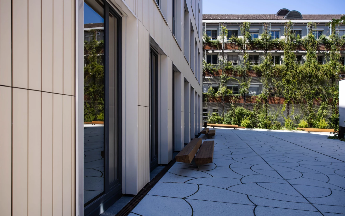 A modern building facade with vertical panels and large windows, next to a courtyard with patterned grey tiles and wooden benches. A (Baerahus, Feldkirch)