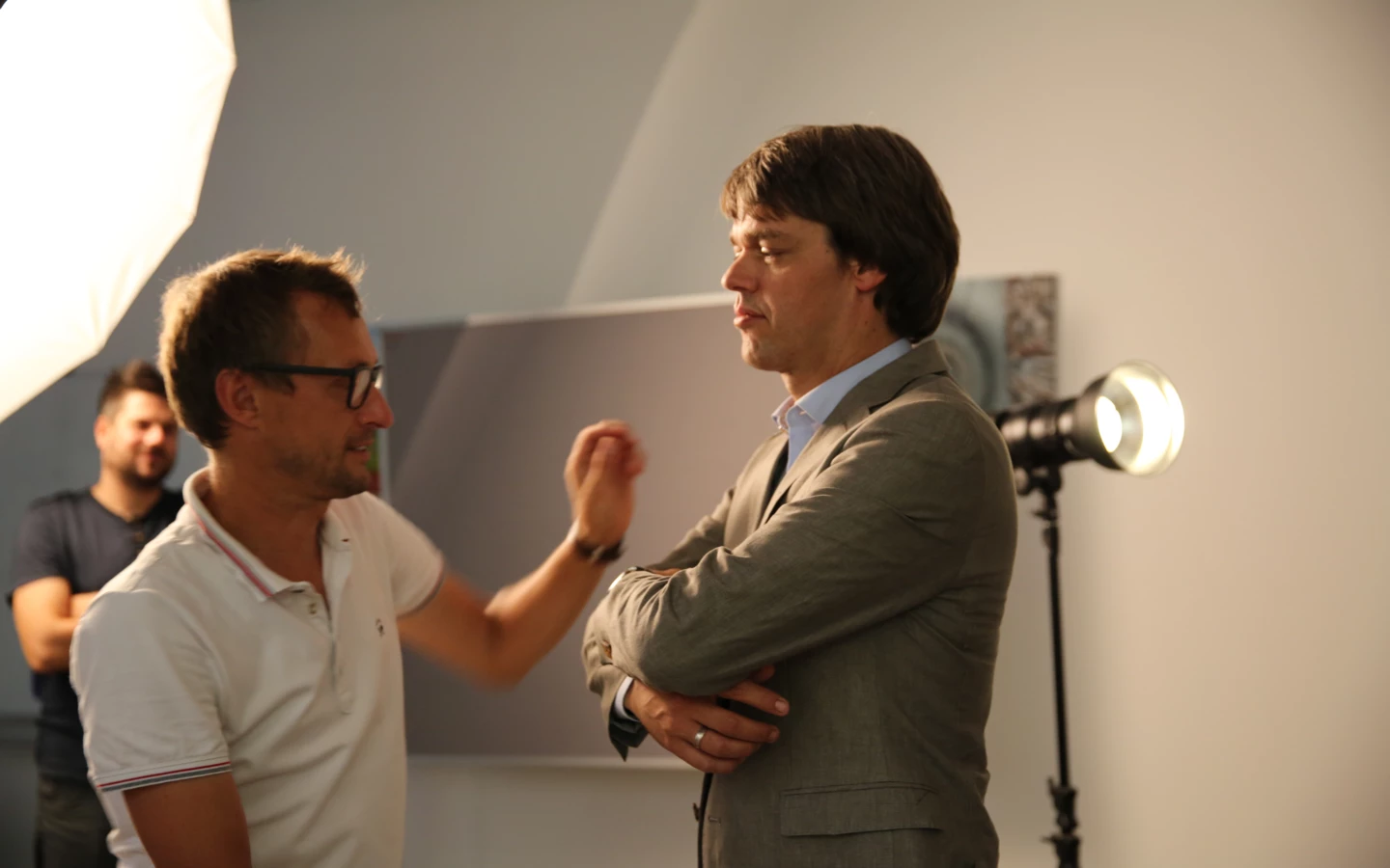 Zwei Männer in einem Studio mit Beleuchtung, die sich unterhalten. Ein Mann im Anzug hat die Arme verschränkt.