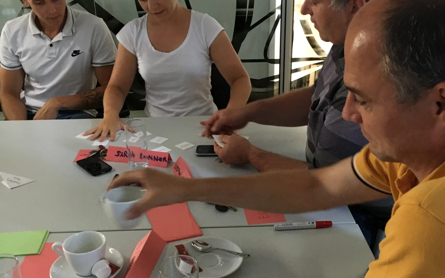 Teilnehmer eines Workshops sitzen an einem Tisch, diskutieren und ordnen Karten mit Notizen. Kaffeetassen und Wassergläser stehen auf dem Tisch.