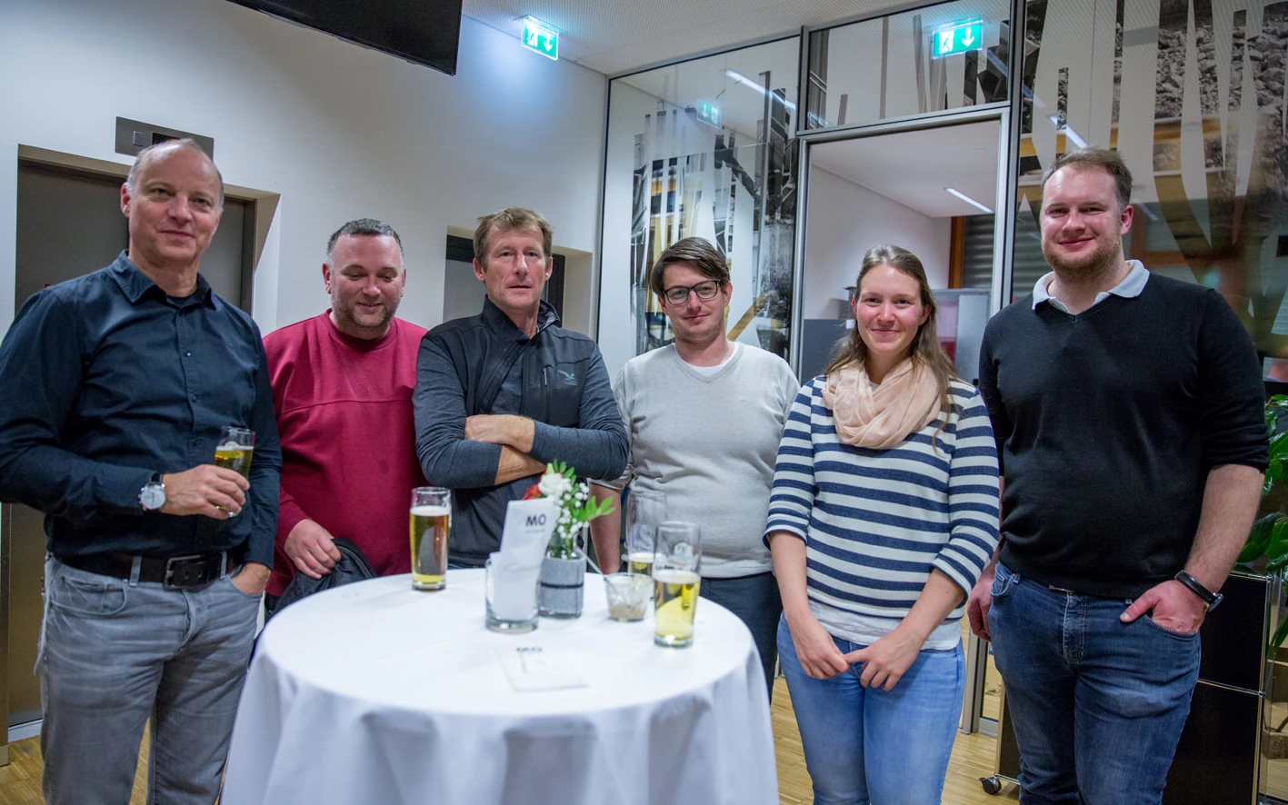 Eine Gruppe von sechs Personen, drei Frauen und drei Männern, stehen lächelnd bei einem Event mit Getränken auf einem Stehtisch (Hubert, Geburtstag).